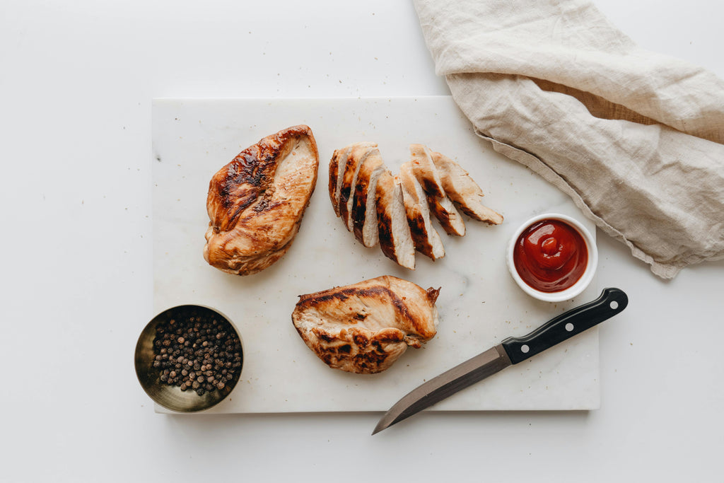Sliced chicken breast on a marble cutting board with a knife, small bowl of sauce, and pepper grinder.